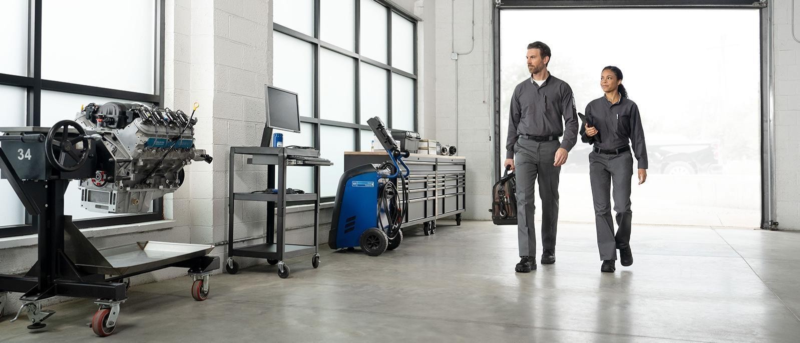 Two automotive technicians walking through a clean service bay with tools, equipment, and an engine display in a modern auto repair facility.